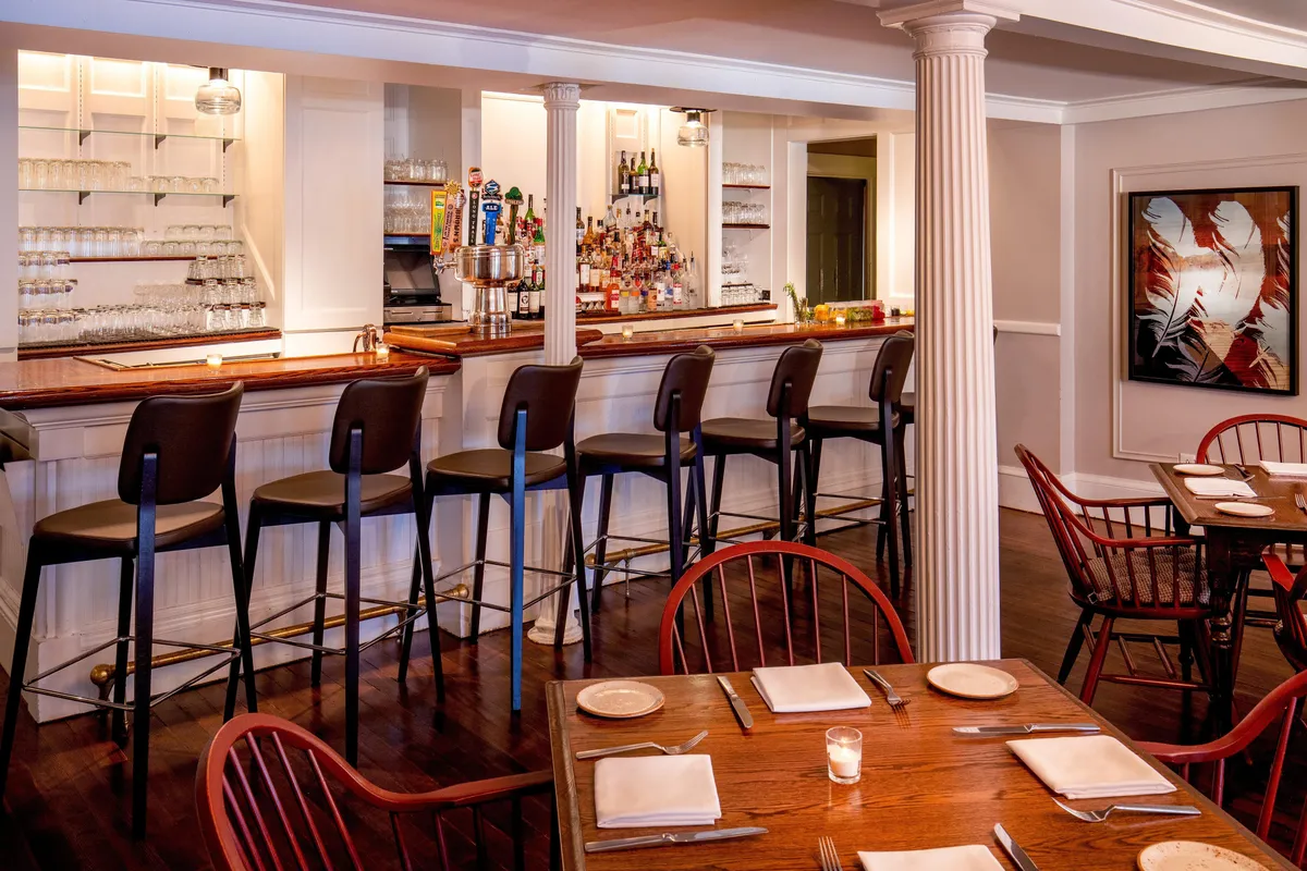The image features a stylish bar area with comfortable stools lined up along a wooden counter, surrounded by wooden dining tables and adorned with local artwork on the wall. The atmosphere is lively with guests enjoying drinks, sharing plates of gourmet food, and engaging in animated conversations.