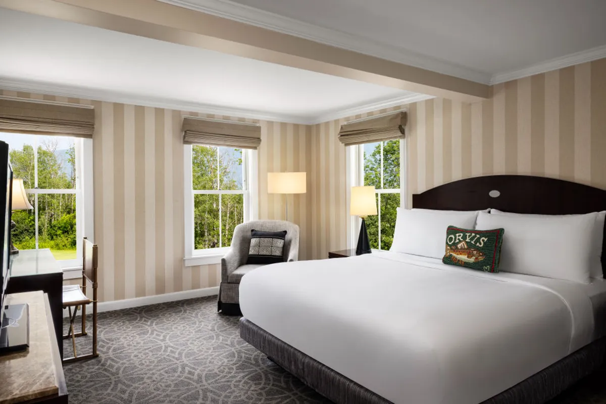 A beautifully appointed suite bedroom featuring a plush king-sized bed with custom bedding, a cozy armchair next to large windows, and a television. Natural light fills the room, creating a warm and inviting atmosphere that highlights both the classic and contemporary touches.