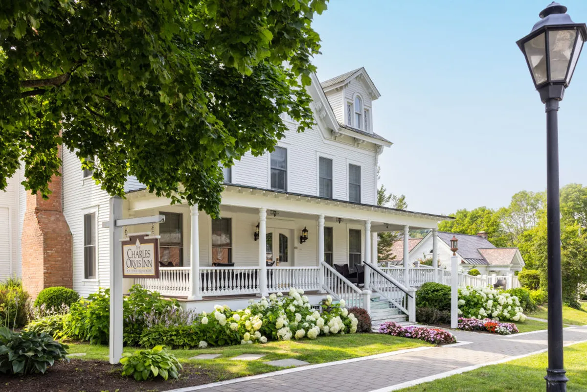 An exterior shot of an ornate New England-style porch adorned with lush flower beds, thoughtful signage, and a classic lamp post. The porch exudes a welcoming, serene ambiance, enhanced by morning or evening light and the changing seasons visible in the garden.