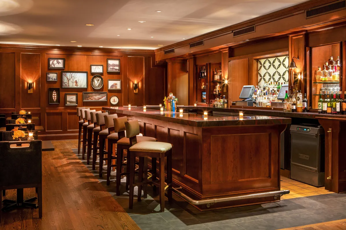This warmly lit interior scene features a sophisticated bar counter with elegant bar stools, shelves of bottles, and tasteful wall art. Guests are seen socializing and relaxing, highlighting a refined atmosphere suited for wine or scotch tasting by the fire pit.