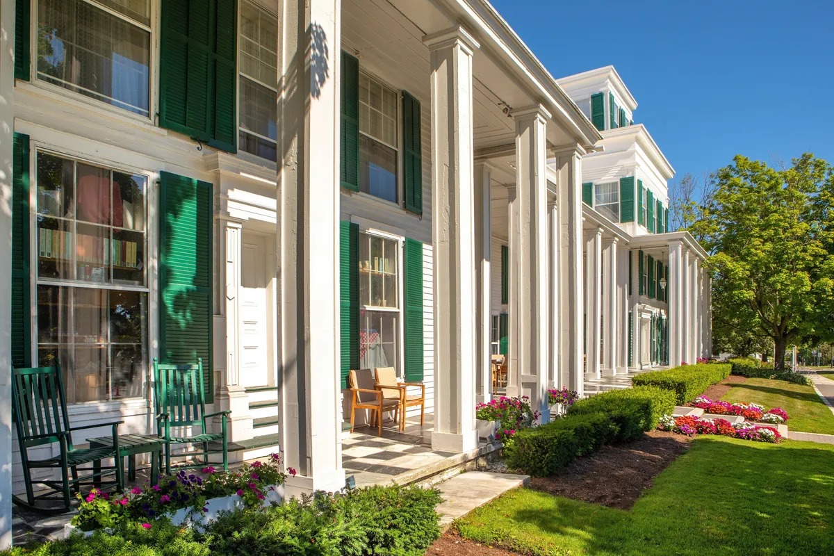 A tranquil porch scene with white columns, green shutters, and inviting rocking chairs nestled among colorful flower beds. The setting is perfect for reading, enjoying a morning coffee, or relaxing with a pet beside you. The peaceful garden and soft light reinforce the sense of welcome and respite.