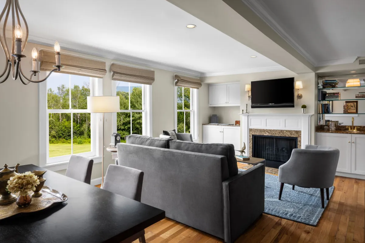 The image showcases a warm and elegant interior with large windows filling the room with natural Vermont light. The inviting fireplace is the focal point, framed by a plush sofa and a stylish dining table, creating a perfect setting for relaxing, entertaining, or simply enjoying the serene surroundings.