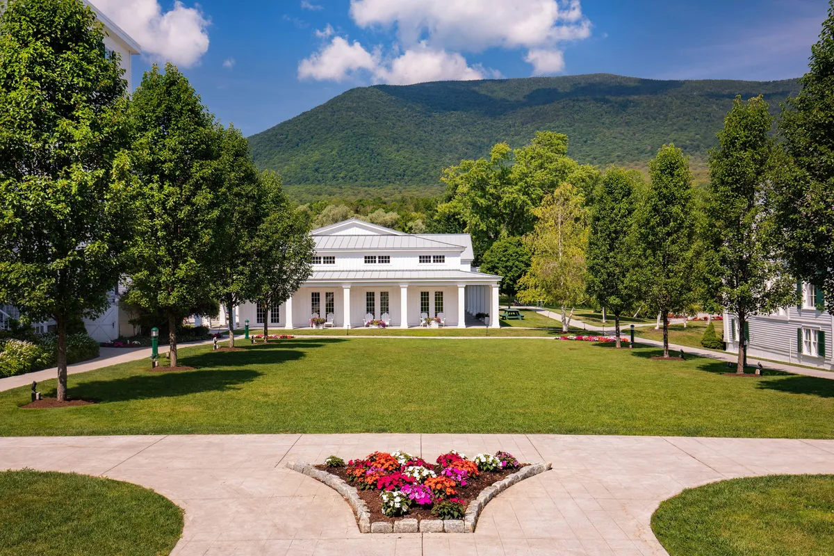 This image captures the main building of the property with a welcoming porch, a vibrant flower bed in the foreground, and a scenic mountain backdrop. The well-maintained lawns are visible, suggesting ample outdoor space for activities and relaxation.