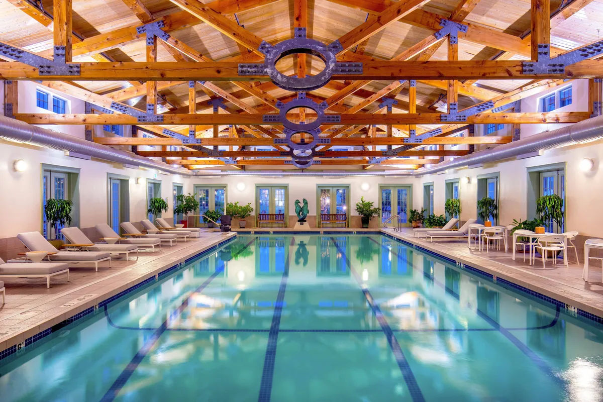 A serene indoor swimming pool surrounded by wooden beams, lounge chairs, decorative plants, and a cozy seating area, inviting guests to relax, swim, or gather by the pool.